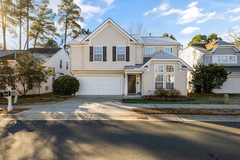 Suburban beige house with a two-car garage and wreath on the door. Sunny day, trees in the background, and shadows on the driveway.