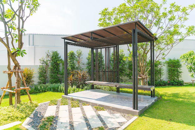 Modern pergola with a wooden bench on a stone patio in a lush garden. Green grass, trees, and shrubs create a tranquil atmosphere.