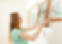 Woman in a green shirt hangs floral paintings on a beige wall using a hammer. The room appears bright and orderly, evoking a calm mood.