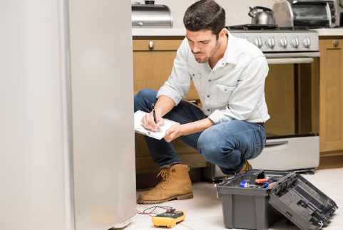 Man kneeling in a kitchen, taking notes on a clipboard near a fridge. Open toolbox on the floor suggests appliance repair. Neutral tones.