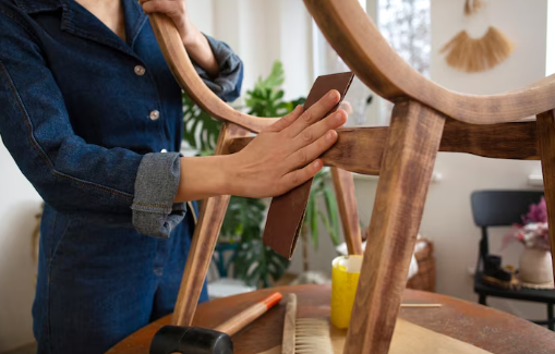 Person sanding a wooden chair indoors, wearing denim. Tools and plant in background. Bright, natural lighting creates a focused, calm mood.