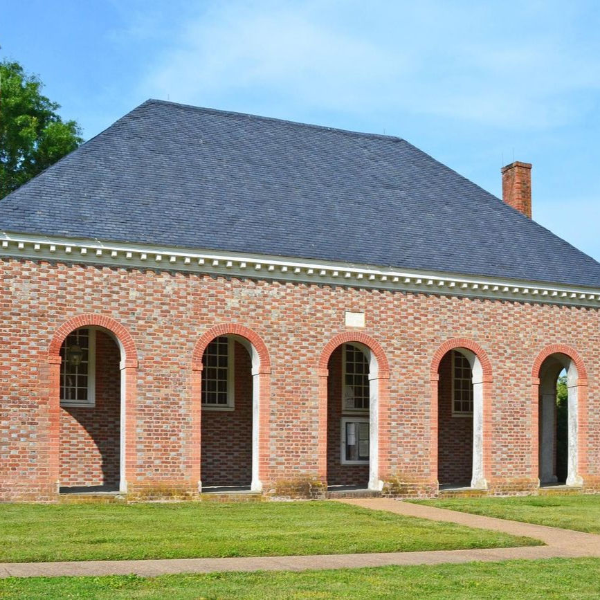 Brick building with arched windows, blue roof, and chimney. Grass and walkway in the foreground. Clear blue sky background.