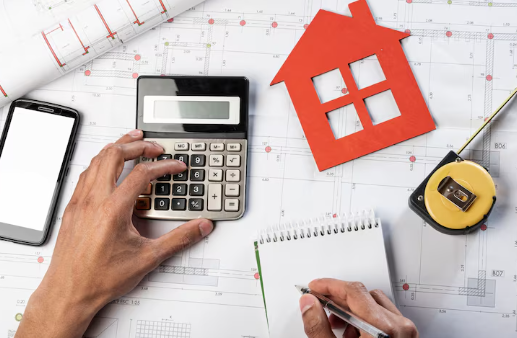 Hands using a calculator and notebook on architectural plans. A red house cutout, smartphone, and tape measure are visible.