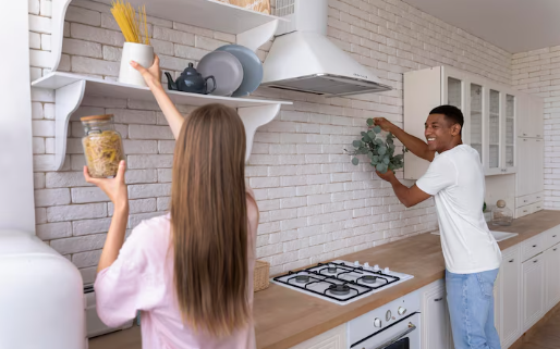 A woman reaches for pasta on a shelf while a man places a plant in a bright kitchen with white brick walls, creating a cheerful atmosphere.