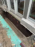 Basement waterproofing work near a set of glass doors. Brown dimple board installed along the foundation. Visible green tarp and tools.