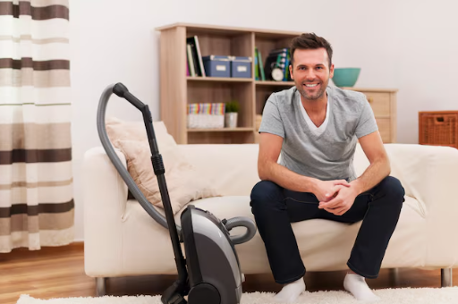 Man smiling, sitting on a couch next to a vacuum cleaner in a cozy living room. Neutral tones, striped curtains, books on shelves behind.