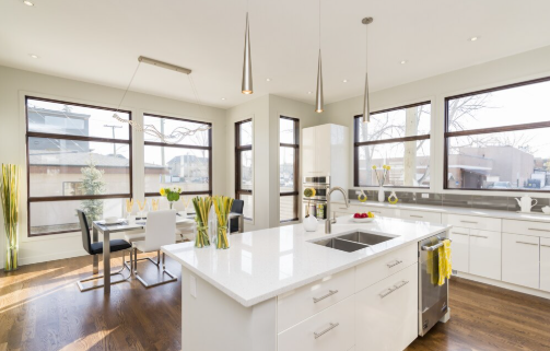 Bright modern kitchen with white cabinets, wooden floor, and a central island. Large windows, pendant lights, and a small dining table.
