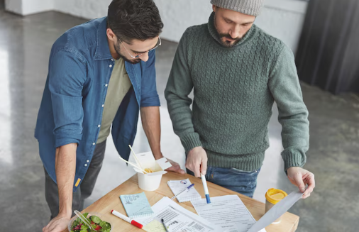Two men review documents on a table with food containers and pens in a bright room. One points at a paper; both appear focused.