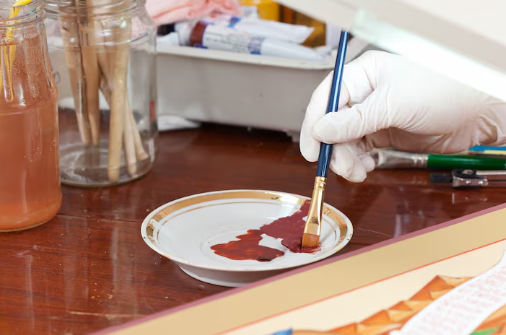 A gloved hand paints on a plate with a brush. Paint tubes and glasses sit on a wooden table. The scene is well-lit and focused.