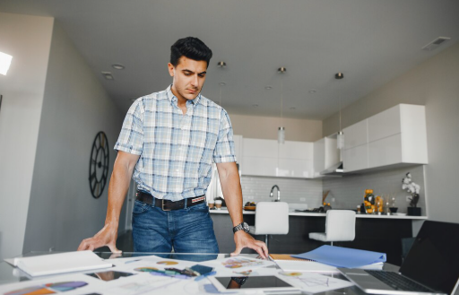 Man in plaid shirt examines documents on a table in a modern kitchen. Neutral tones, bright lighting, laptop open nearby.