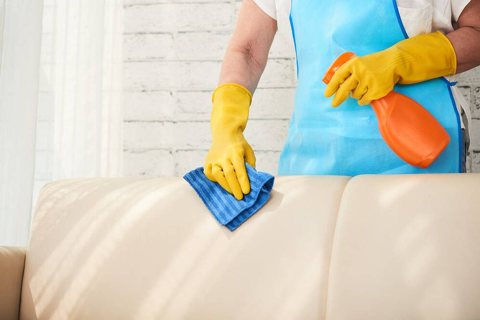 Person in blue apron and yellow gloves cleaning a beige sofa with a spray bottle and blue cloth against a white brick wall background.