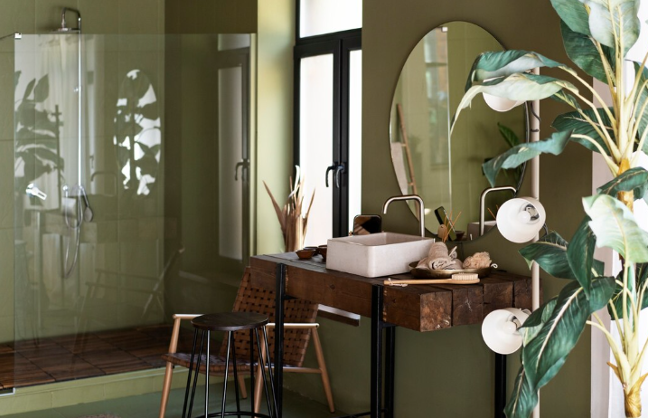 Bathroom with a wooden vanity, oval mirror, and green walls. A large plant nearby and a glass shower in the background create a serene vibe.
