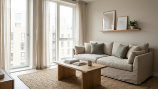 Cozy living room with beige sofa, green cushions, wooden coffee table, and rug. Art and plant on a wall shelf. Large windows with sheer curtains.