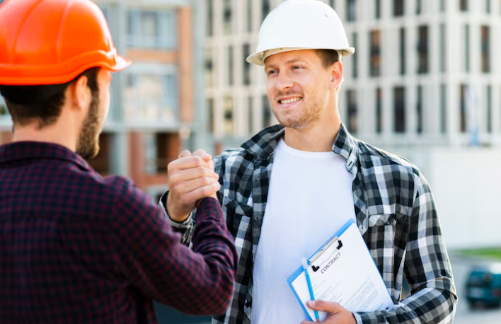 Two construction workers in hard hats shake hands at a building site. One holds a clipboard labeled "Contract," both appear pleased.