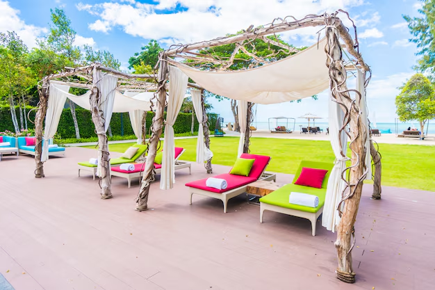 Wooden cabanas with white drapes and colorful sunbeds on a deck overlooking a green lawn and blue sky, creating a relaxing atmosphere.