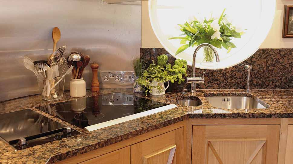 Cozy kitchen with a granite countertop, induction stove, wooden cabinets, and round window. Vase of flowers and plants add freshness.