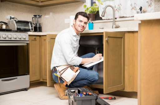 Man kneeling in a kitchen checking under a sink, holding a clipboard. Toolbelt and toolbox nearby. Bright setting, light brown cabinets.