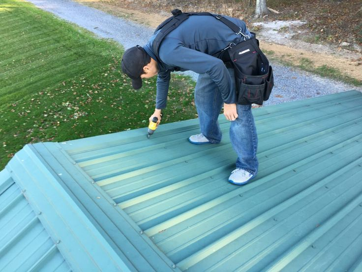 A person in blue jeans and a cap uses a drill on a green metal roof. They're wearing a black tool belt. Green grass and gravel path in the background.