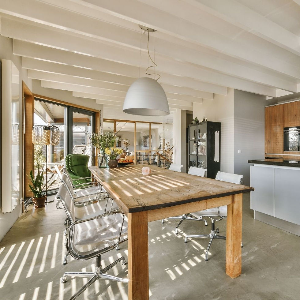 Modern dining room with a wooden table, white chairs, and a green armchair. Light streams through large windows, casting shadows. Relaxed vibe.