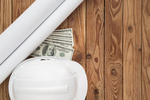 White hard hat, rolled blueprints, and dollar bills on a wooden surface. The scene suggests construction or architectural planning.