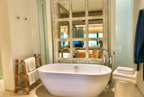 Modern bathroom with a white freestanding tub, large decorative mirror, towel rack, and neatly folded towels. Bright, serene ambiance.