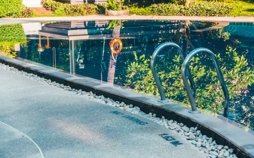 Poolside view with metal ladder, clear water, and a life preserver. Greenery reflects in the water. "No Diving 1.6M" text on the edge.