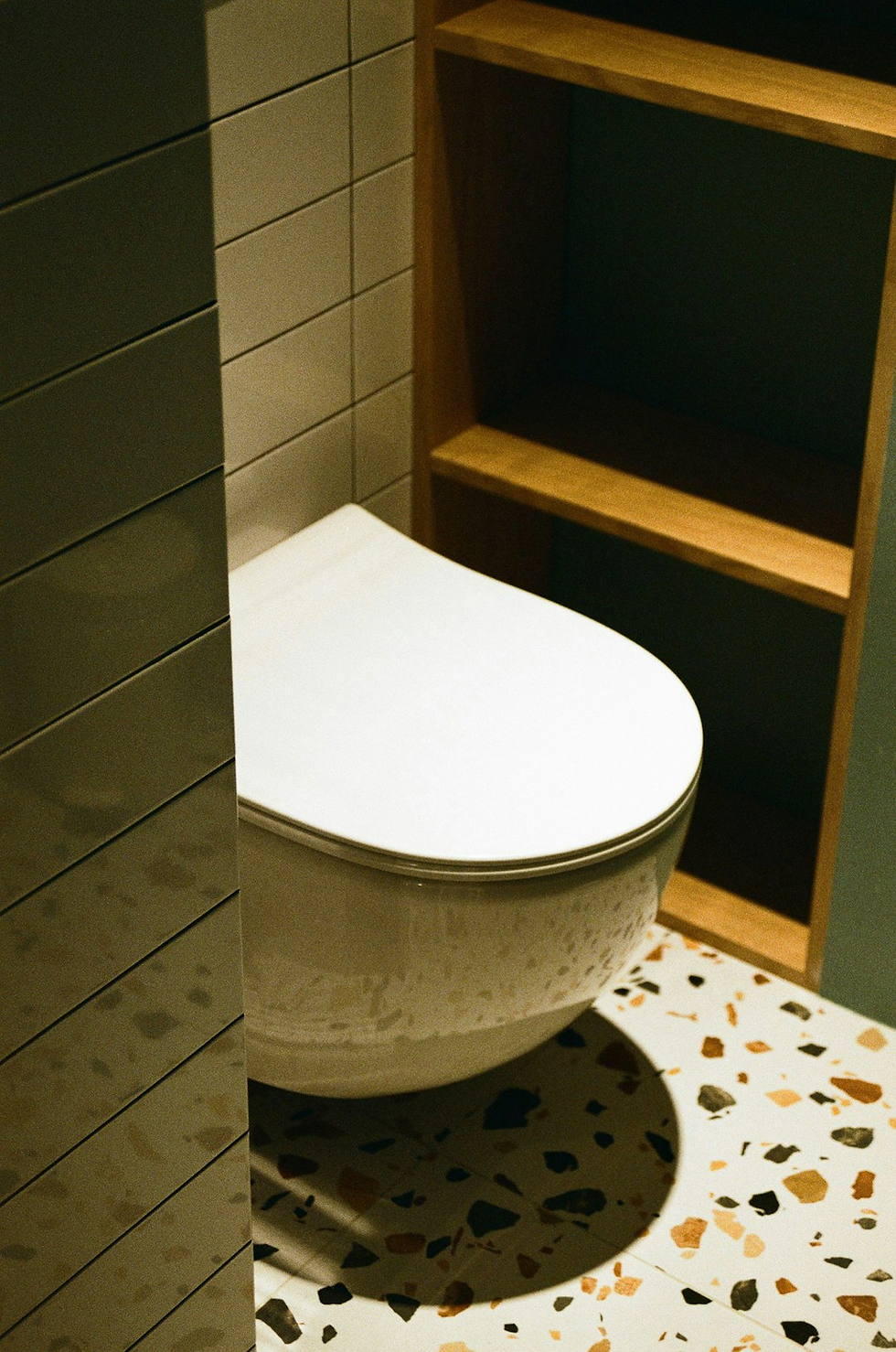 Modern white toilet with closed lid beside a wooden shelf in a bathroom. Terrazzo floor with speckled pattern, tiled walls.