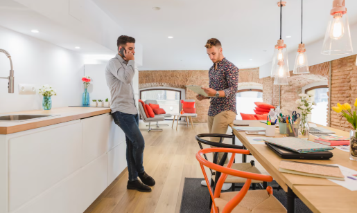 Two men in a modern office with brick walls; one on the phone, the other reading a document. Bright, airy setting with red chairs.