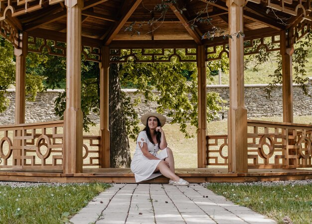 Woman in a white dress and hat sits in a wooden gazebo, with trees and a stone wall in the background, creating a serene atmosphere.