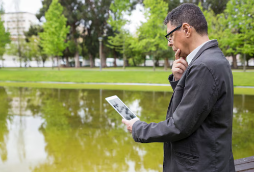 Man in glasses reads tablet by pond, wearing black jacket. Green trees and grass reflect in water, creating a serene atmosphere.