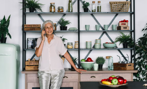 Elderly woman talks on phone, smiling in a modern kitchen with a black shelf, potted plants, and vibrant fruit. Text reads "ENJOY YOUR LIFE."