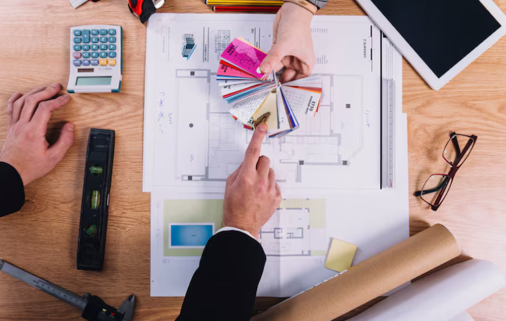 Hands point at color swatches on blueprints, surrounded by a calculator, tablet, and tools on a wooden table, suggesting design planning.