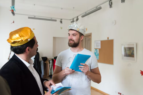 Two men in costume crowns hold blue folders, conversing in a bright room with modern decor and coats in the background.