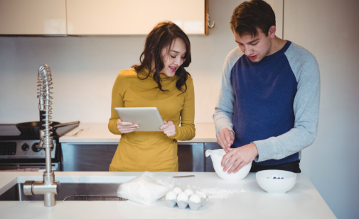A man and woman in a kitchen, the woman holds a tablet while the man mixes ingredients in a bowl. Eggs and flour are on the counter.