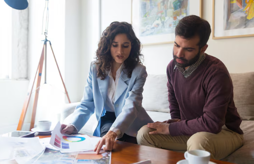 Two people in discussion, sitting on a sofa with documents on a table. Bright room, art on walls, focused expressions, casual attire.