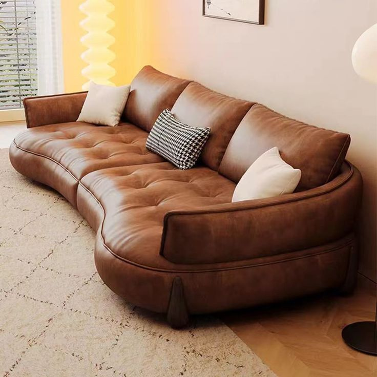 Curved brown leather sofa with white and black houndstooth pillows. Set in a warmly lit room with a beige rug and framed artwork. Cozy mood.