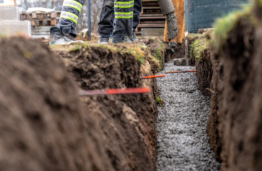 Construction worker pouring concrete into a trench. Worker wears boots and reflective pants. Surrounding are soil mounds and red markers.