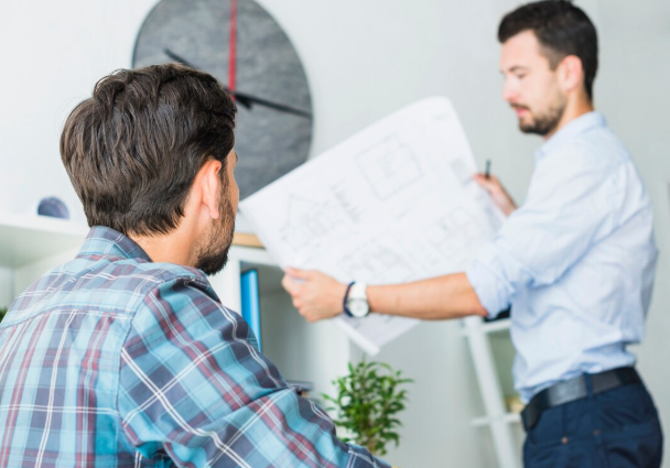 Two men in a bright room; one in plaid listens while the other in blue presents a blueprint. Modern clock on the wall, plants nearby.