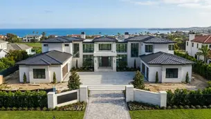 Luxurious modern mansion with dark roofs and large windows, sits by the ocean. Palm trees line the property, evoking a serene coastal vibe.