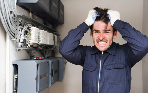 Man in blue coveralls holds head in frustration, standing by electrical panels with cables. Wearing gloves, he shows a tense expression.