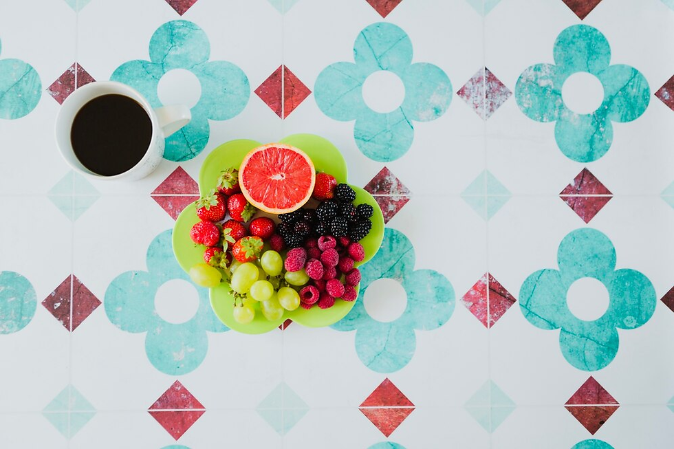 Cup of coffee beside a green plate with assorted fruits on a patterned turquoise and red table. Bright, fresh, and inviting scene.