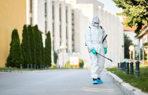 Person in hazmat suit sprays disinfectant on a street near a building, with neatly trimmed hedges beside. Bright, sunny day.