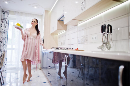 Woman in pink robe smiling in modern kitchen, holding dish with fruit. Bright, spacious setting with white cabinets and knives in holder.