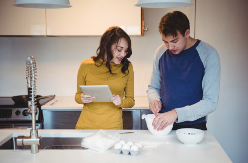 Young couple baking in a modern kitchen; woman in a yellow top holds a tablet, man in a blue shirt mixes ingredients. Eggs and flour nearby.