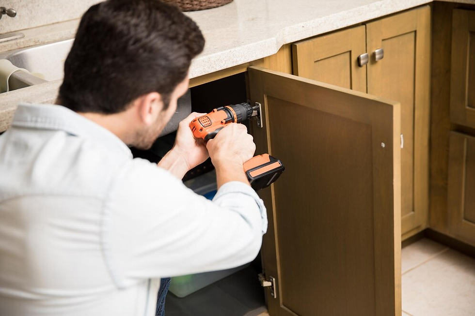 Man uses an orange drill to fix a wooden cabinet under a kitchen sink. The setting is bright and tidy, with soft brown and beige tones.