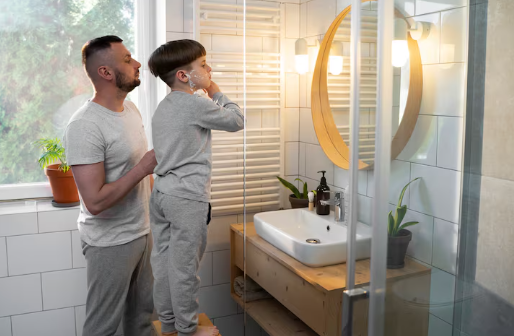 Boy standing on a stool, shaving cream on face, assisted by man in gray pajamas in a bathroom with plants and round mirror, warm lighting.