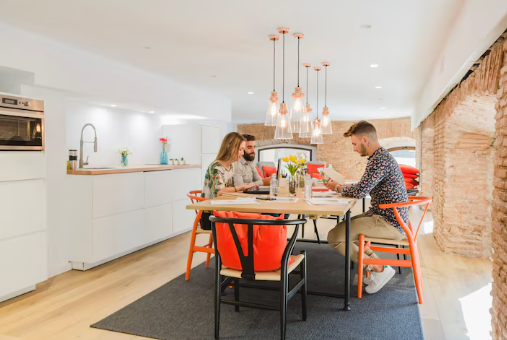 Three people sit at a table with laptops and papers in a bright kitchen with brick walls. Pendant lights and orange chairs add color.