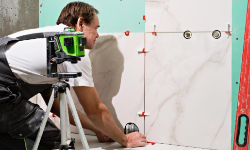 Worker in overalls installing white marble tiles with laser level on tripod. Red spacers and leveler visible. Bright interior setting.
