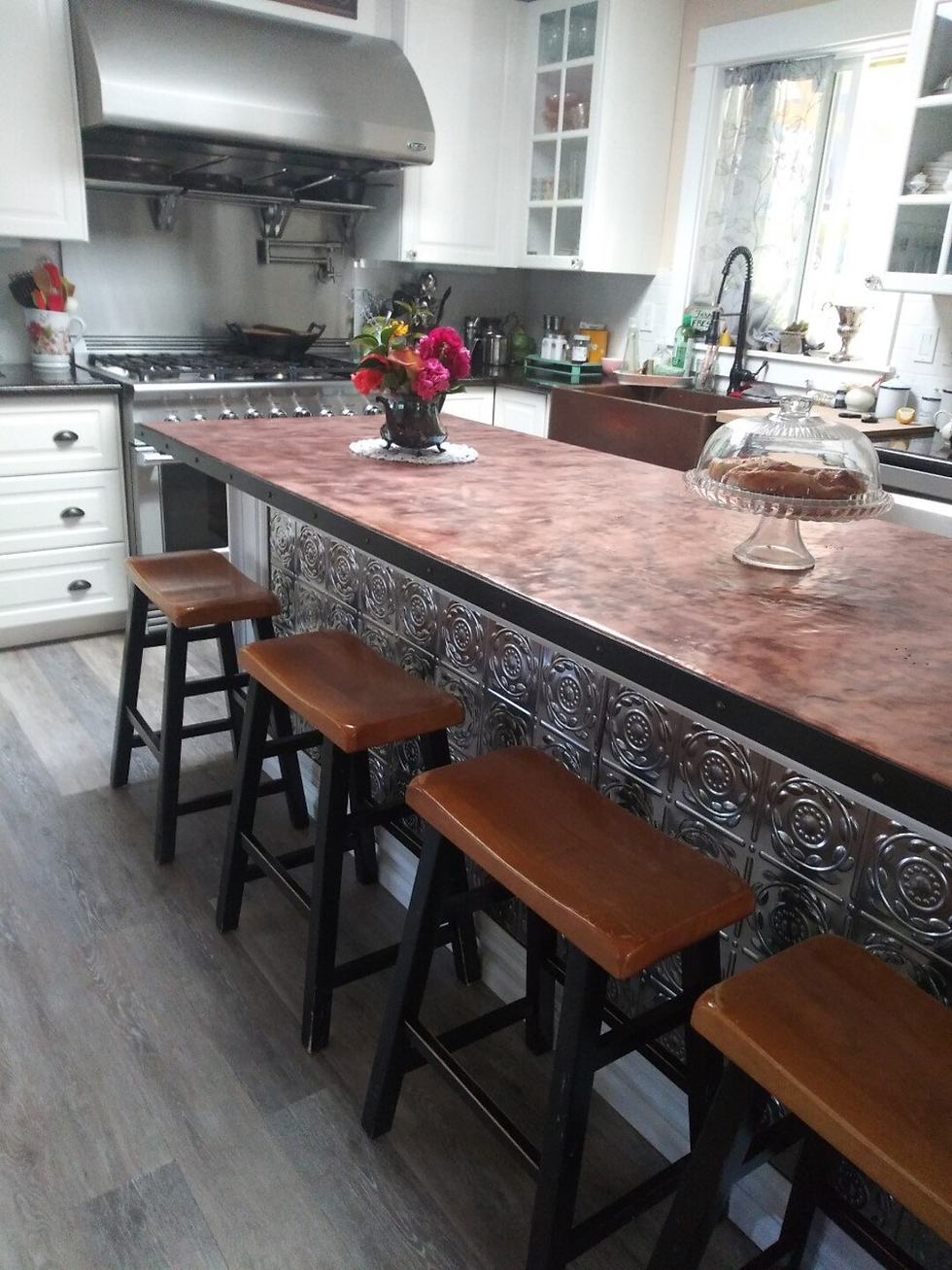 Kitchen with a brown island, four stools, flowers in a vase, and a cake under a glass dome. Stainless steel appliances and white cabinets.