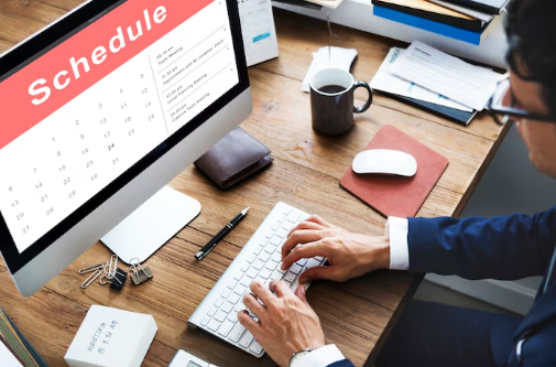 Person in a suit typing at a desk with a computer displaying a "Schedule" calendar. Nearby are a coffee cup and notepad. Office setting.